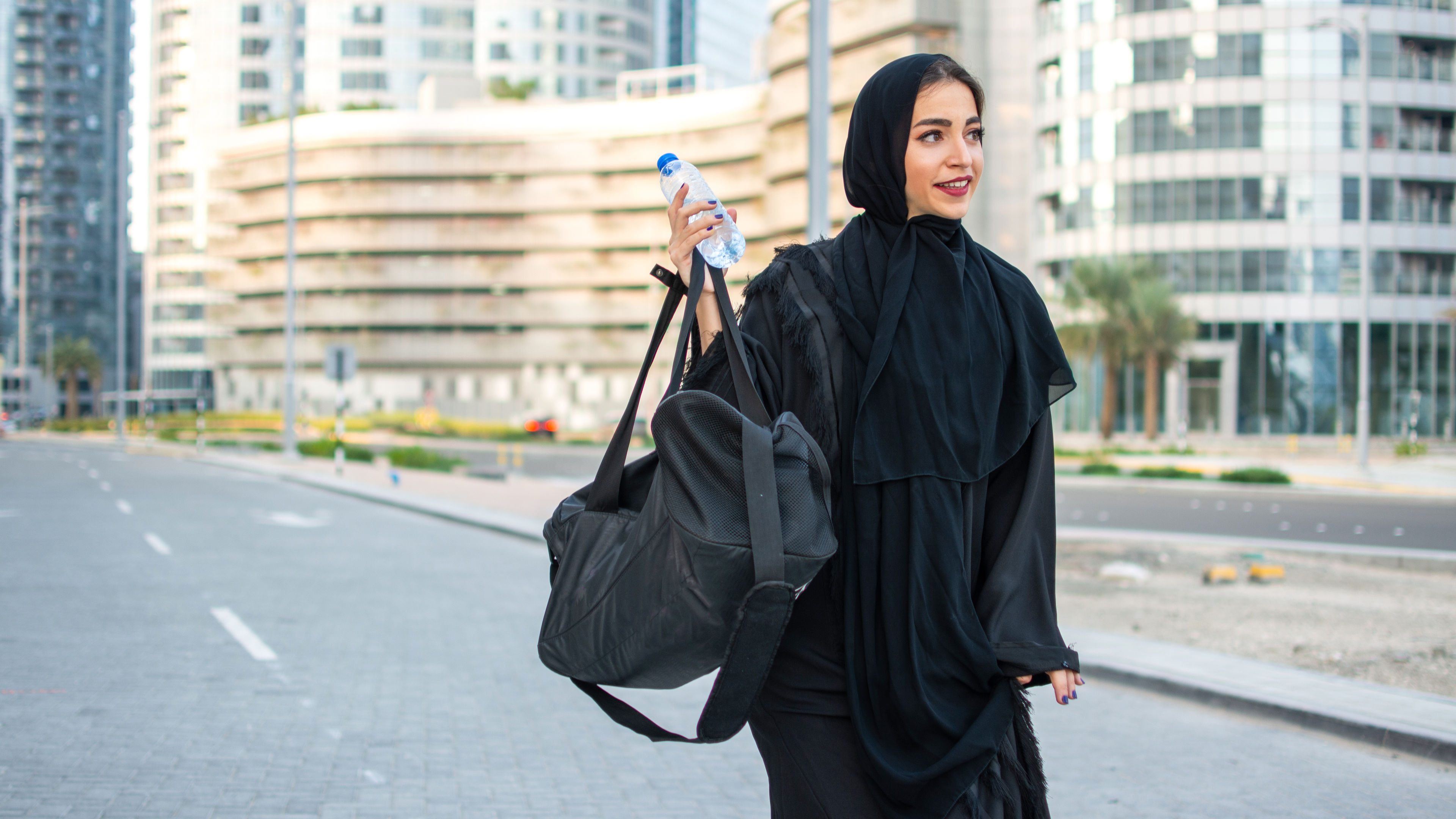 Beautiful Arab woman in abaya holding water bottle and a sport's bag on her way to a gym