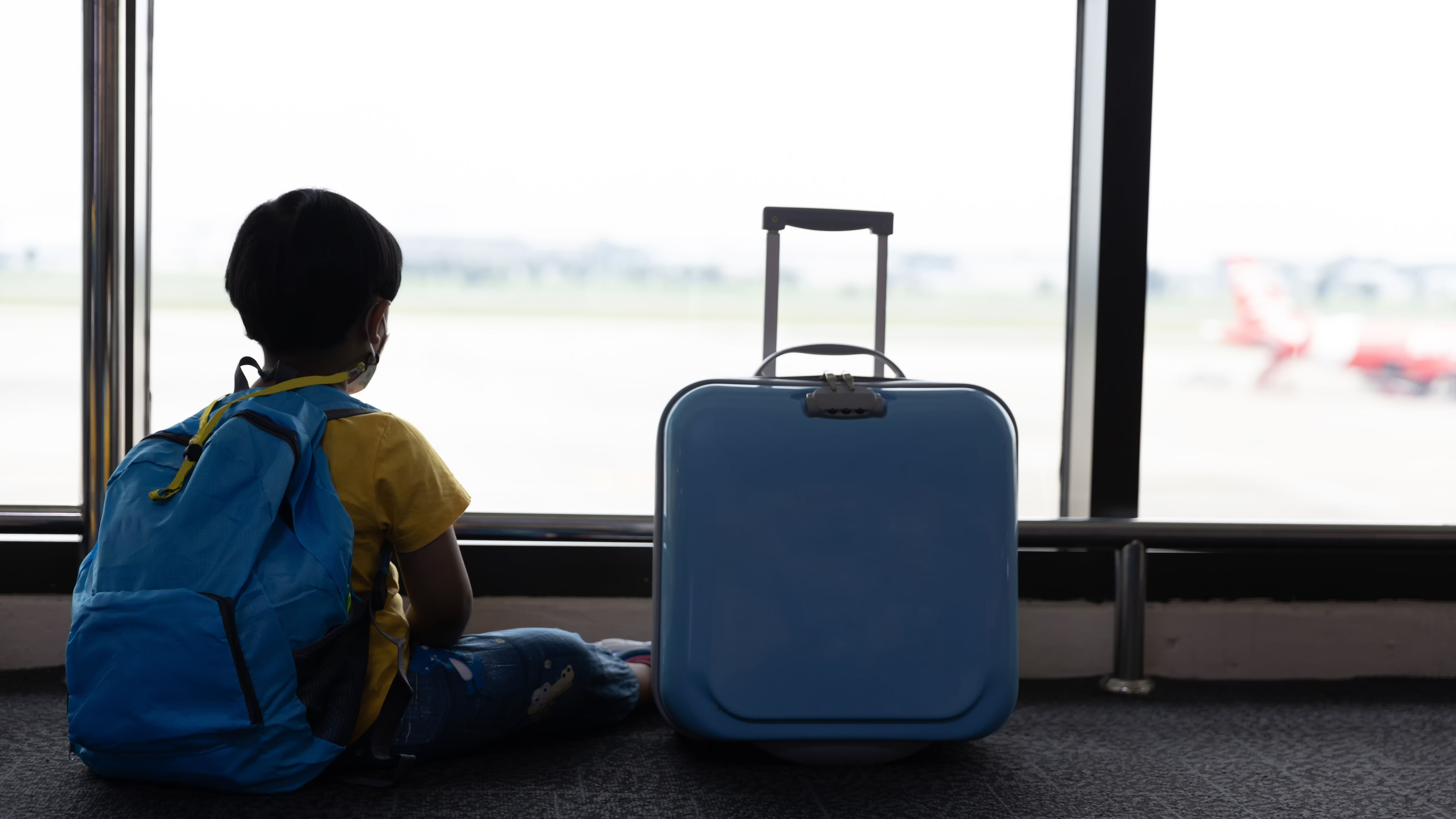 Child with suitcase at airport terminal waiting for departure looking out the window.