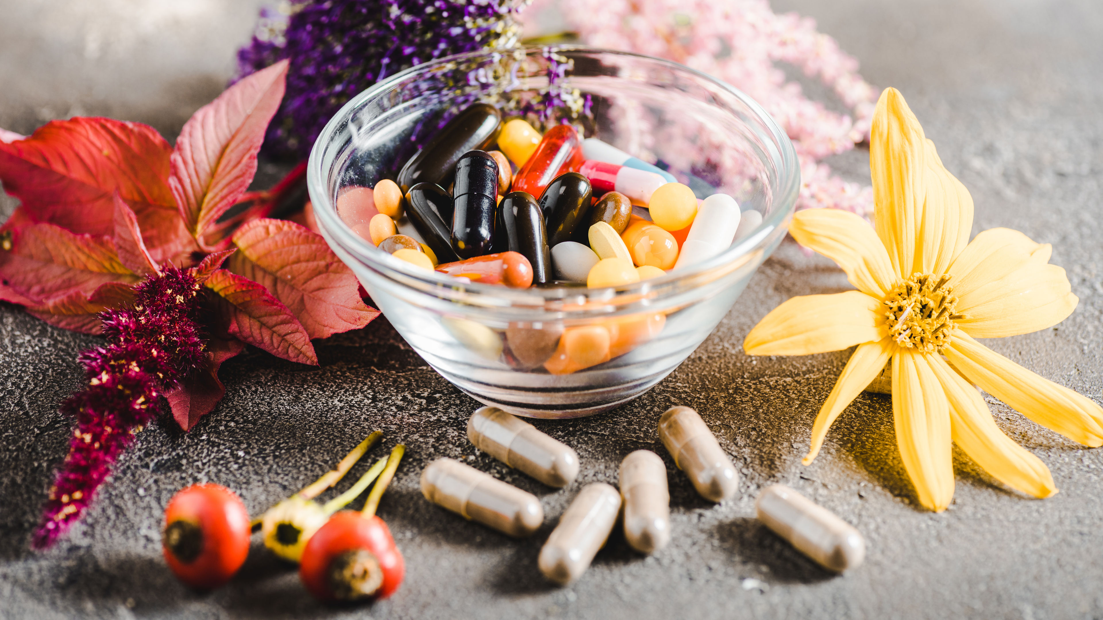 Pharmacological pills in glass bowl and colored flowers on wooden tabletop, alternative medicine