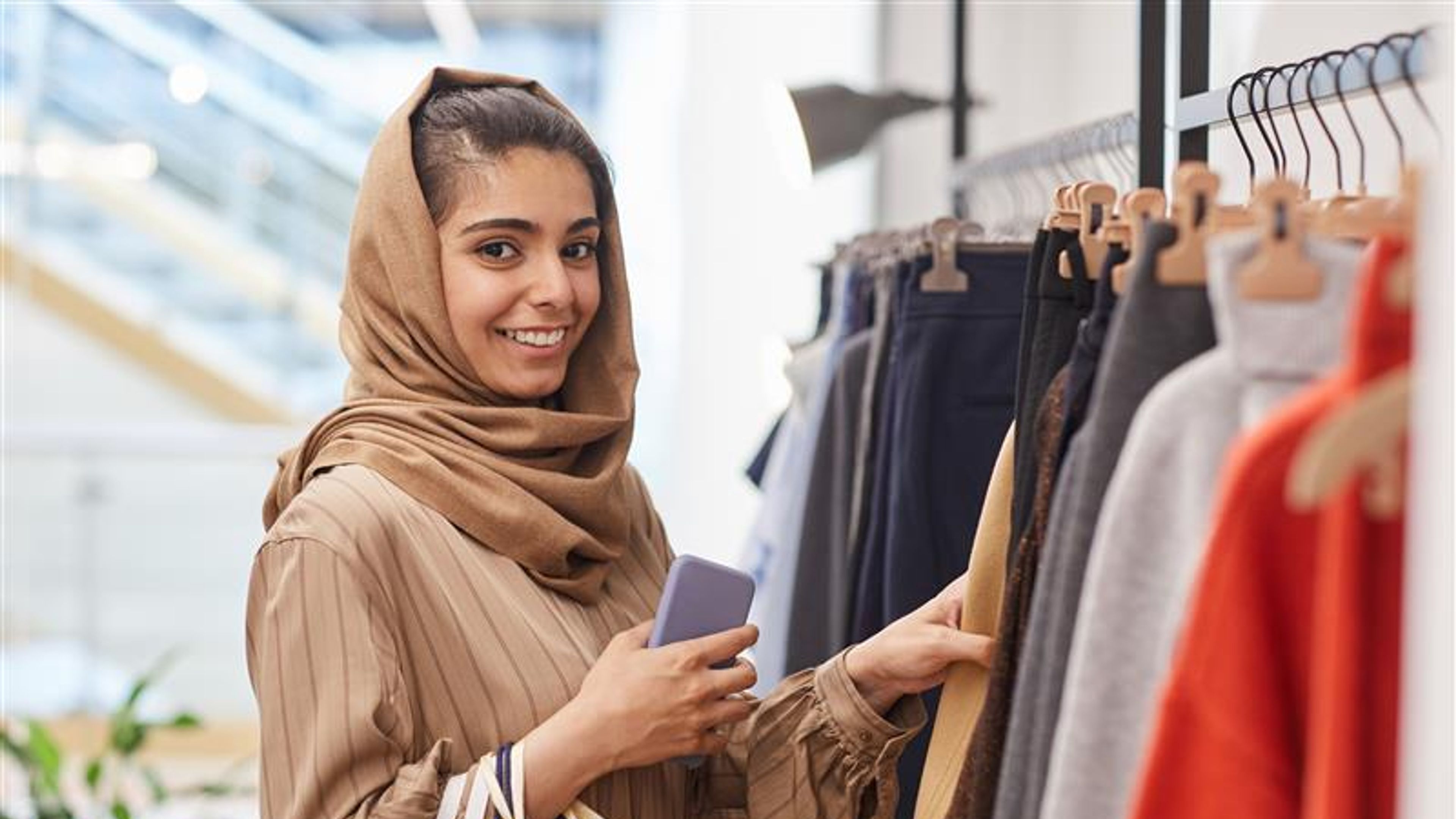 Waist up portrait of elegant young woman looking at camera while standing by clothing racks and enjoying shopping in mall. 